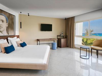 Helles Hotelzimmer mit großem Bett, Meerblick, Wandbild, Fernseher und gemütlicher Sitzgruppe.