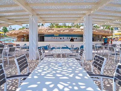 Terrasse eines Hotels mit weißen Möbeln und gemustertem Schatten unter einem Holzvordach.