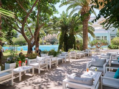Terrasse mit weißen Sitzmöbeln, Bäumen und Palmen neben einem blauen Pool unter Sonne.