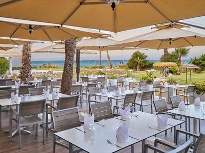 Freiluftrestaurant mit Tischen und Stühlen unter großen Sonnenschirmen, Blick auf Meer und Palmen.