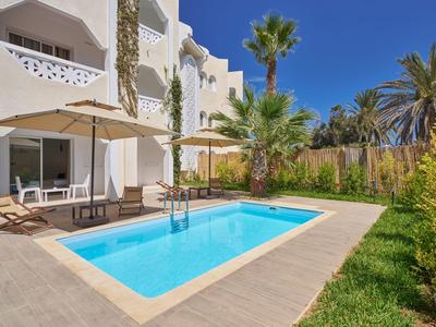 Moderner Pool mit blauem Wasser, Sonnenliegen, Schirmen und weißen Gebäuden bei klarem Himmel.