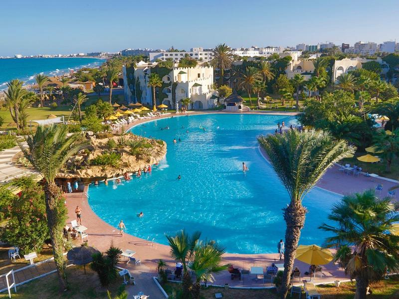 Großer Hotelpool mit Palmen und Sonnenschirmen in tropischer Umgebung am Meer.