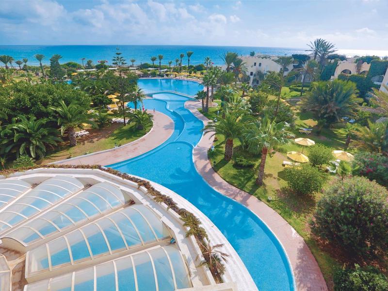 Hotel mit tropischem Garten, Pool und Meerblick im Hintergrund.