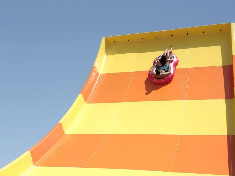 Person fährt auf rotem Schlauchboot eine große, gelb-orange Rutsche hinunter bei klarem Himmel.