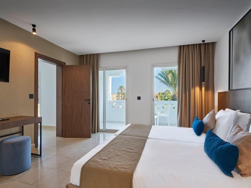 Helles Hotelzimmer mit großem Bett, Fernseher und Blick auf eine Terrasse mit Meerblick.