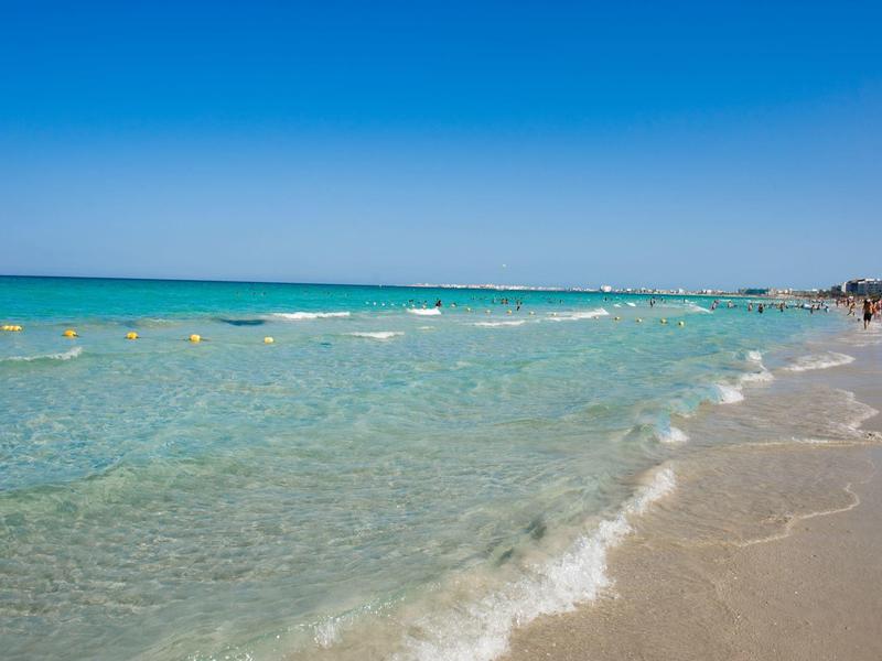 Klares Meer mit sanften Wellen an einem sonnigen Strand unter blauem Himmel.