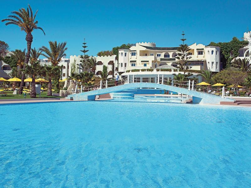 Großer Poolbereich mit Brücke, umgeben von Palmen und einem Hotelgebäude bei klarem Himmel.