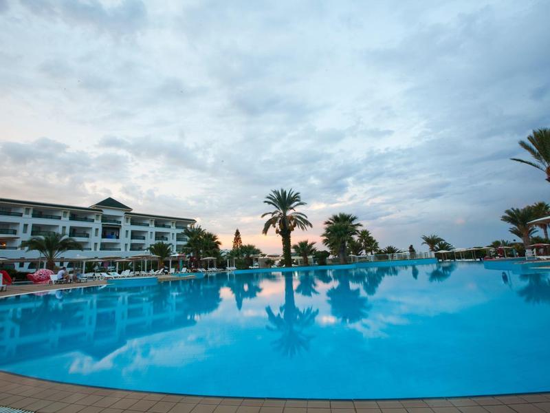 Large outdoor pool in front of a hotel building with palm trees under a cloudy sky.