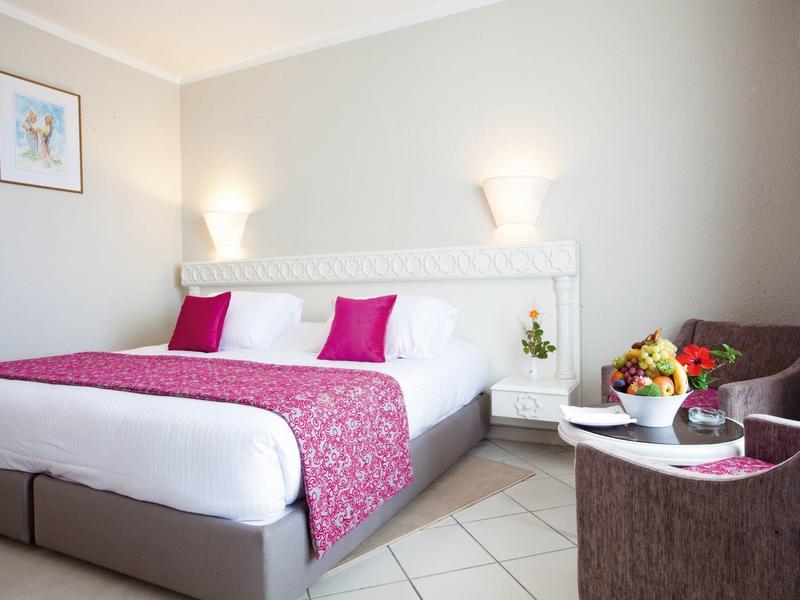 Modern hotel room with double bed, white walls, pink accents, and a small table with fruit.