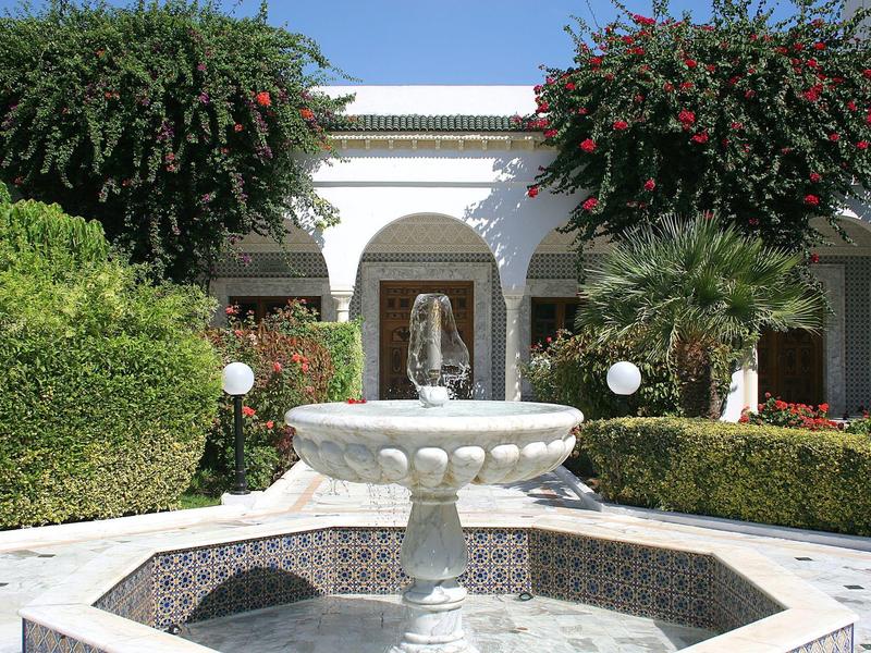 Courtyard with fountain and statue, surrounded by green plants and white building.