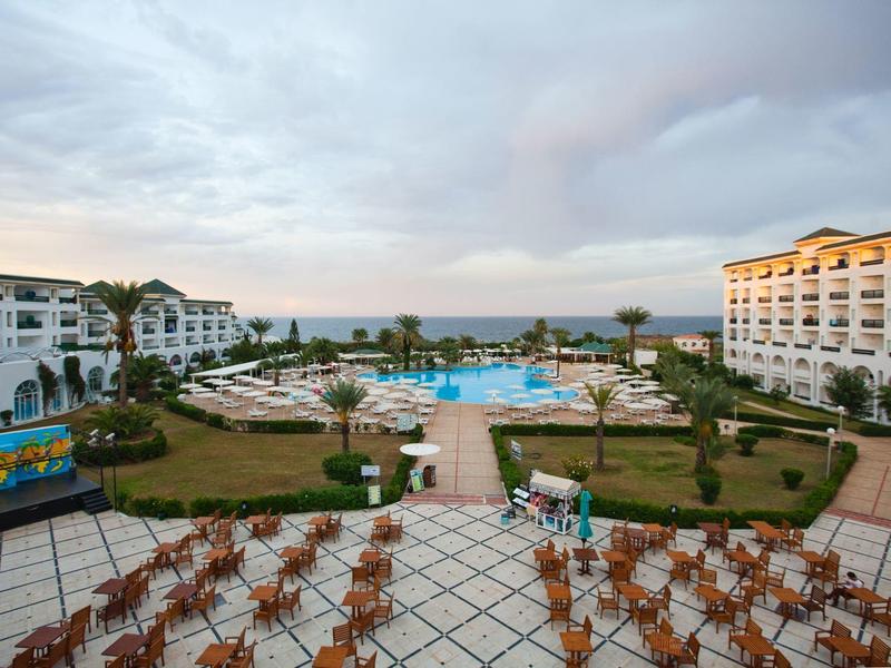 View of a resort with pool, sun loungers, and two hotels under a cloudy sky.