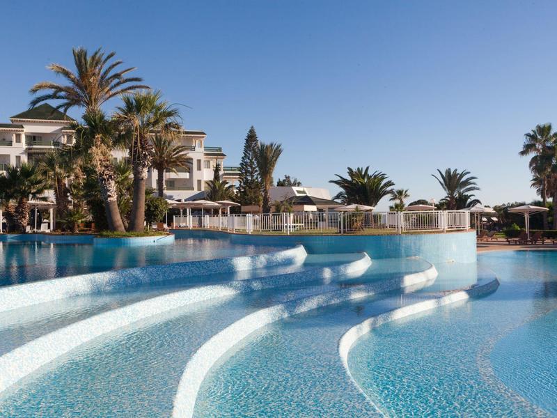 Large hotel pool with white steps and bar stools in the water under a clear sky.
