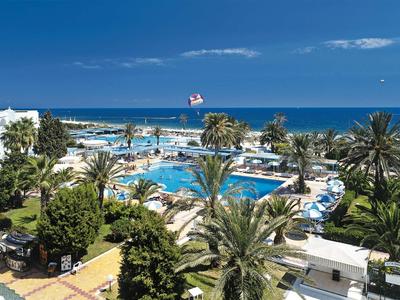 Große Poolanlage mit Palmen und Liegestühlen, umgeben von weißen Gebäuden und Blick auf das blaue Meer.