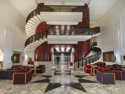Grand salon d'hôtel avec double escalier en colimaçon et sièges modernes aux couleurs chaudes.