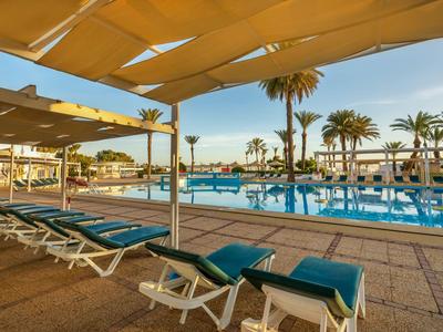 View of a pool area with lounge chairs under sunshades and palm trees at sunset.