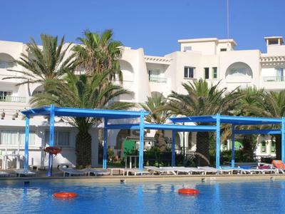 Blaues Schwimmbecken vor weißem Hotelgebäude mit Palmen und Sonnenschirmen.