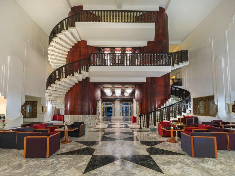 Grand salon d'hôtel avec double escalier en colimaçon et sièges modernes aux couleurs chaudes.