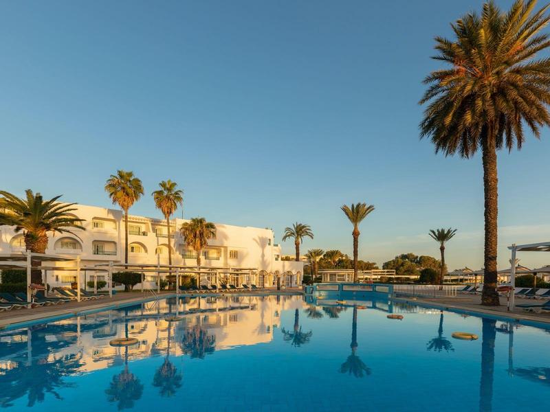 Grand piscine extérieure devant des hôtels blancs avec des palmiers et un ciel clair au coucher du soleil