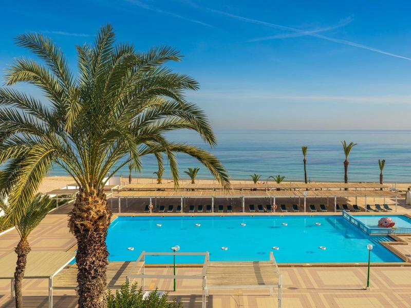 Grande piscine avec des palmiers et vue sur la mer calme sous un ciel clair.
