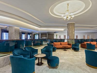 Bright hotel lobby with modern blue armchairs, orange sofas, and marble columns.