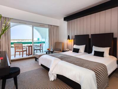 Modern hotel room with two beds, balcony access with sea view, and seating area.