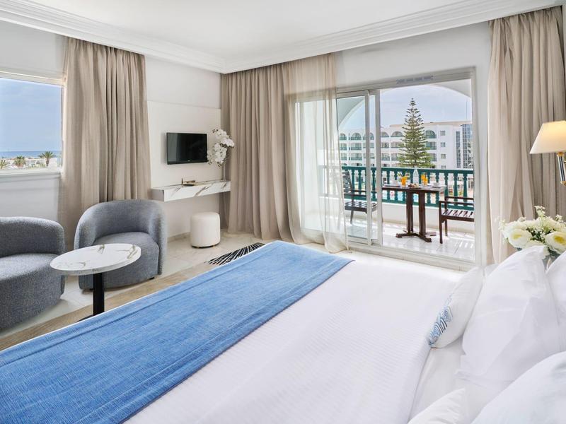 Bright hotel room with double bed, two chairs, balcony, and sea view.