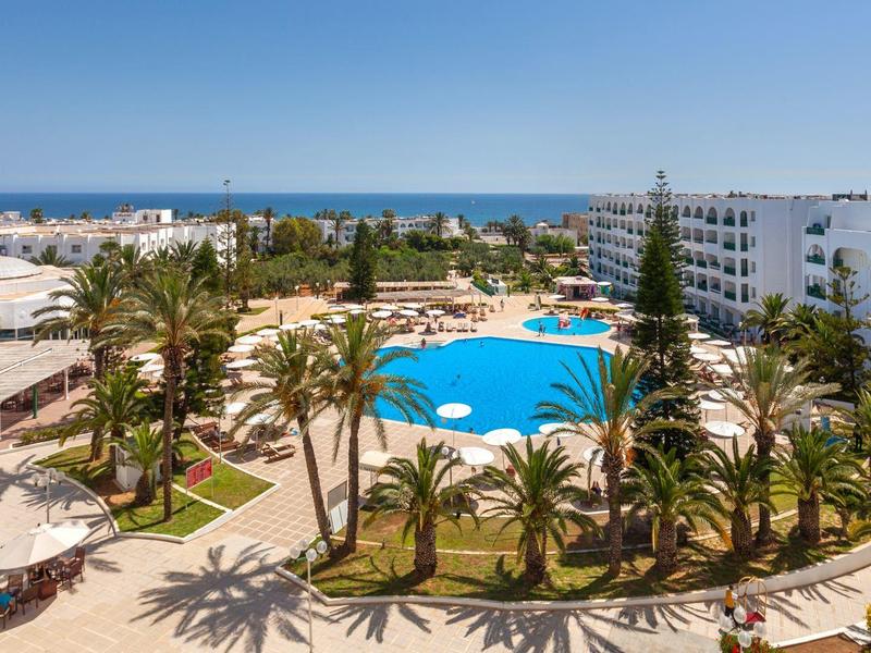 Large hotel pool surrounded by palm trees and white buildings with sea view.