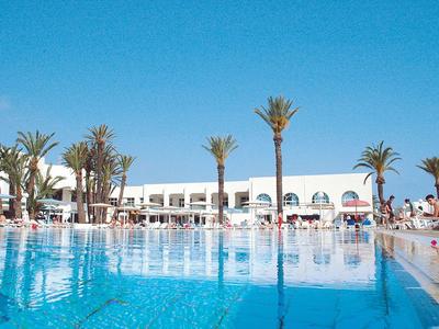 Grande piscine avec eau bleue claire devant un hôtel blanc et des palmiers.