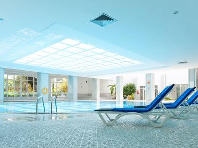 Piscina coperta luminosa con lettini blu e grandi finestre che lasciano entrare luce naturale.