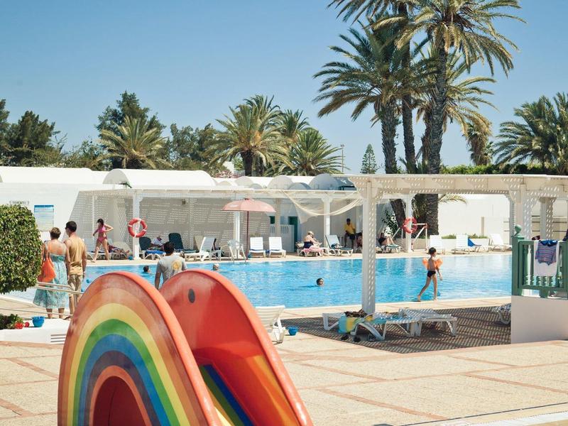 Scivolo d'acqua arcobaleno colorato accanto a una piscina all'aperto con palme sotto un cielo blu.