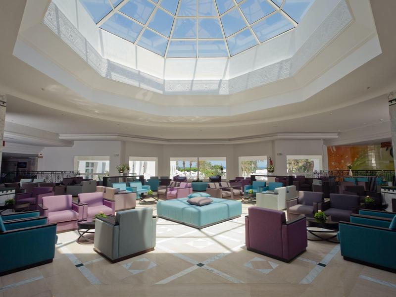 Hall dell'hotel luminoso con cupola di vetro, sedute moderne blu e viola e luce naturale.