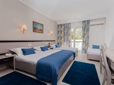Bright hotel room with double bed, blue bedspread, chair, and balcony window with curtains.