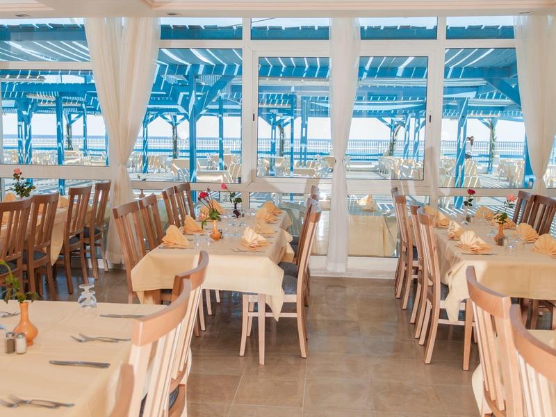 Bright restaurant with wooden chairs and tables overlooking a terrace and the sea.