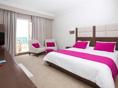 Camera d'hotel moderna con letto grande, accenti viola e zona salotto vicino alla finestra.