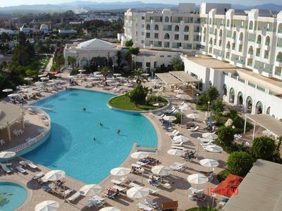 Grande hotel con piscina curva e numerosi lettini all'aperto.