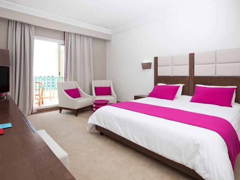 Camera d'hotel moderna con letto grande, accenti viola e zona salotto vicino alla finestra.