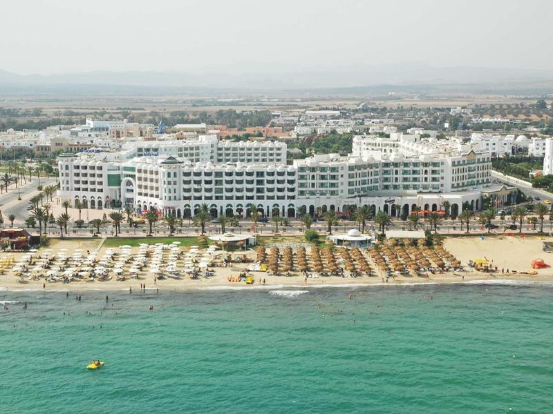 Grande hotel sulla spiaggia con lettini e acqua di mare limpida in primo piano.