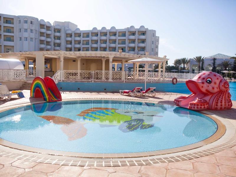 Piscina per bambini con giochi d'acqua colorati accanto a un grande edificio dell'hotel sotto un cielo sereno.
