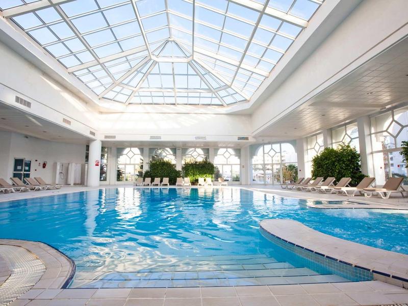 Piscina coperta luminosa con tetto in vetro e sdraio ai lati.