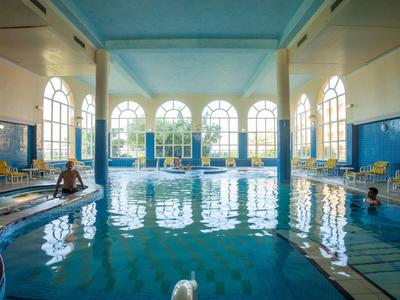 Gran piscina cubierta con agua azul, grandes ventanas y tumbonas amarillas.