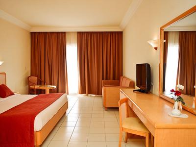 Habitación de hotel luminosa con cama individual, escritorio, silla y grandes ventanas con cortinas.