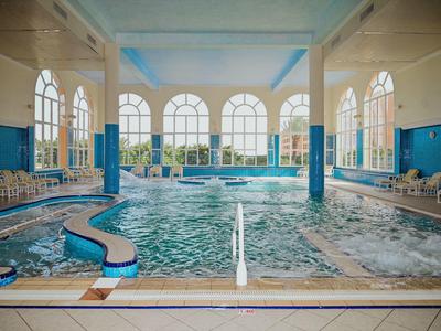 Gran piscina interior con grandes ventanas, sillas blancas y paredes azules en un hotel.