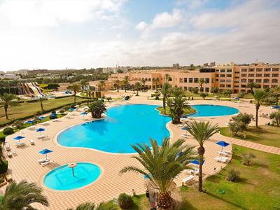 Gran piscina de hotel con tumbonas, palmeras y edificios alrededor bajo cielo nublado.