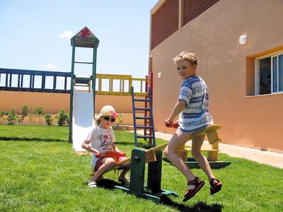 Niños juegan en un balancín junto a un tobogán en un patio con césped junto a una casa.