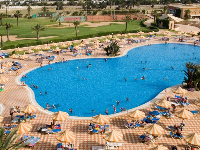 Gran piscina de hotel con muchas tumbonas y huéspedes, rodeada de áreas verdes.