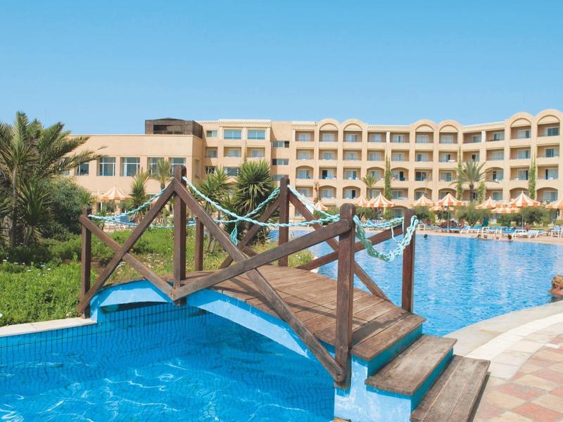 Puente de madera sobre una gran piscina con edificio de hotel al fondo bajo cielo despejado.