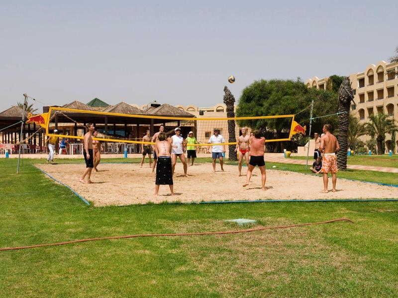 Varias personas juegan voleibol en una cancha de arena junto a un hotel bajo la luz solar.