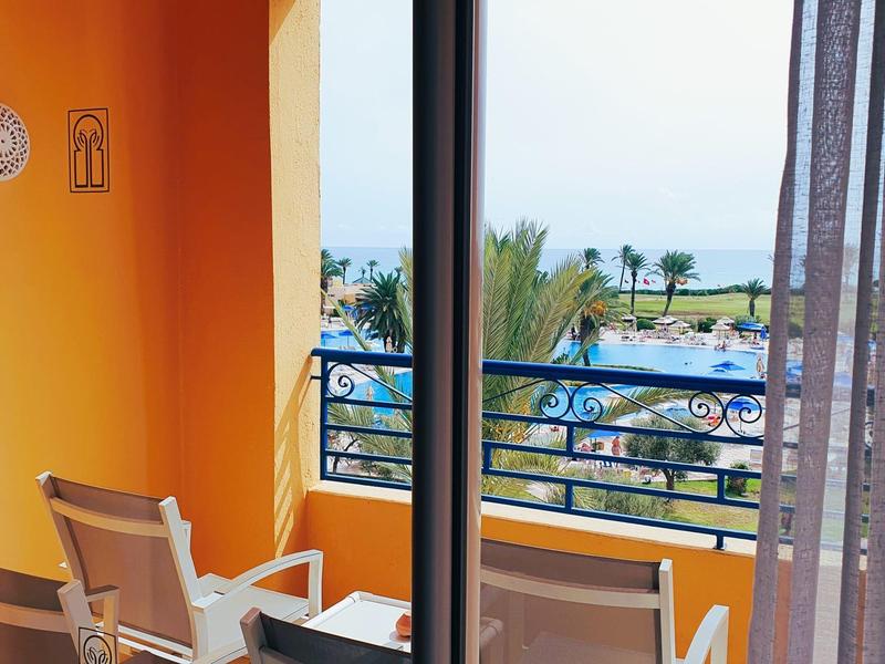 Balcón con sillas, vista a la piscina y al mar bajo un cielo despejado en un hotel.