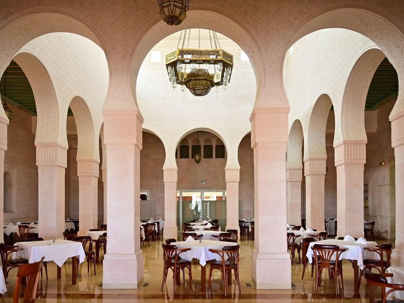 Sala de comedor abierta con altas columnas y arcos redondeados, mesas puestas, iluminación cálida.