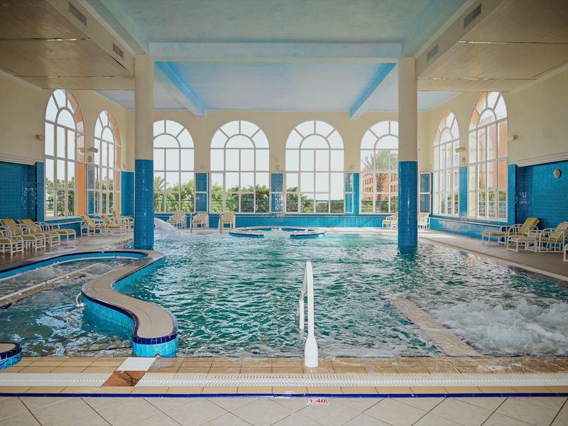 Gran piscina interior con grandes ventanas, sillas blancas y paredes azules en un hotel.